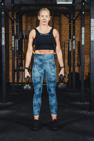 A person wearing mid-rise blue tie-dye leggings and a black sports bra, standing in a gym setting with weights in the background.