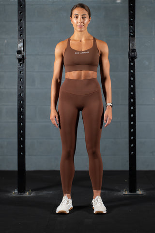 A person wearing chocolate brown high-waisted leggings with a matching sports bra, standing in front of a pair of dark grey exercise bars.